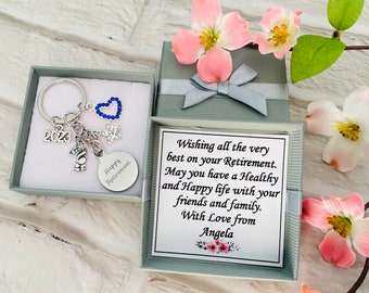 May include: A silver key chain with charms, including a heart with blue rhinestones, a star, a 2023 charm, and a bear charm. The key chain is in a small gray gift box with a white card that reads "Wishing all the very best on your Retirement. May you have a Healthy and Happy life with your friends and family. With Love from Angela."