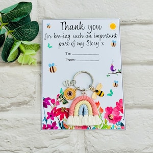 Può includere: Un portachiavi in metallo con un arcobaleno in macramè, un ciondolo a forma di girasole, un ciondolo a forma di ape e un ciondolo a cuore con la scritta Thank You. È fissato su un cartoncino bianco con il testo Thank you for bee-ing such an important part of my Story x.