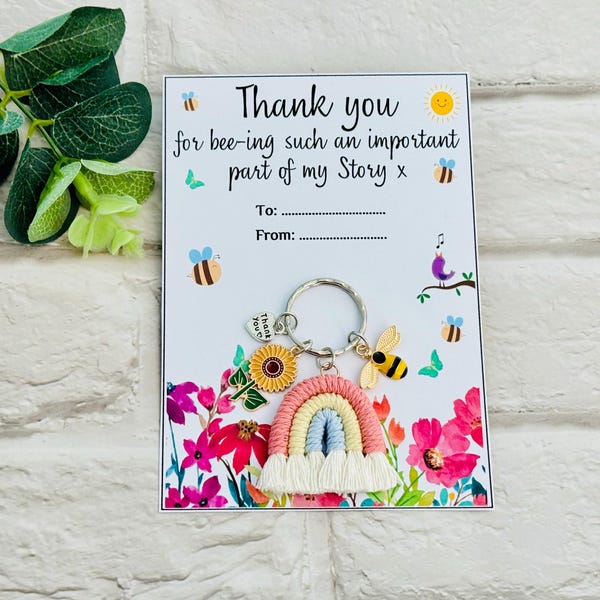 May include: A metal keychain featuring a macrame rainbow, a sunflower charm, a bee charm, and a heart charm with the text Thank You. It is attached to a white card with the text Thank you for bee-ing such an important part of my Story x.