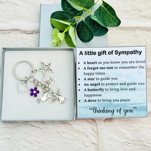 Könnte beinhalten: Ein silberner Schlüsselanhänger mit Anhängern, darunter ein Herz, ein Stern, eine Blume, ein Schmetterling und eine Taube, präsentiert in einer Geschenkbox. Eine Karte mit der Aufschrift "Ein kleines Geschenk des Mitgefühls" mit einer Botschaft des Trostes.