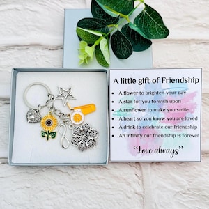 Puede incluir: Un llavero plateado con múltiples dijes, incluyendo un girasol, un corazón, una estrella y un símbolo de infinito, presentado en una caja de regalo. Una tarjeta dice "A little gift of Friendship" con una lista de sentimientos y "Love always."