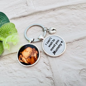 Op de afbeelding: Een zilveren sleutelhanger met twee bedels. Een bedel heeft een zwart-witte echografie van een baby. De andere bedel is een cirkel met de tekst "I can't wait to meet you Mummy" in zwarte tekst met een klein zwart hartje.