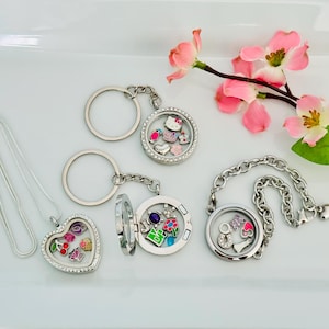 May include: A collection of silver lockets and keychains with various charms. One heart-shaped locket and two round lockets are open, revealing colorful charms. A bracelet with a round locket is also present. Pink flowers with green leaves are in the background.