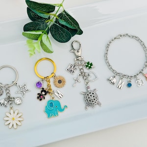 May include: A collection of charm jewelry, including keychains and a bracelet, featuring various charms. Keychains have daisy, star, and elephant charms. The bracelet has letter and number charms, plus a pink flower. The charms are silver and gold-toned.
