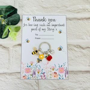 以下が含まれることがあります： ミツバチをテーマにしたキーホルダーが付いたサンキューカード。「Thank you for bee-ing such an important part of my Story」と書かれています。キーホルダーには、ミツバチ、花、リンゴのチャームが付いています。背景には花のイラストが描かれています。