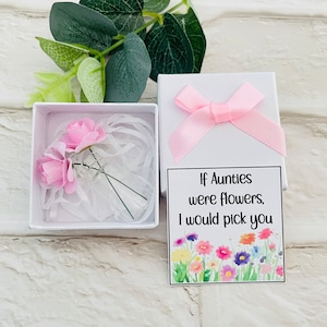 Puede incluir: Una pequeña caja blanca con un lazo rosa en la parte superior. Dentro de la caja hay un pequeño ramo de flores rosas. La caja también incluye una tarjeta con el texto "Si las tías fueran flores, te elegiría" y una ilustración de acuarela de flores coloridas.