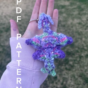 May include: A handmade crochet dragon keychain in shades of purple, green, and pink. The dragon has a detailed design with wings and a tail. The keychain is held up by a silver ring. The text "PDF PATTERN" is written vertically on the left side of the image.