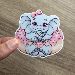 May include: A gray elephant with pink ears and a pink bow sits on a pink donut with purple and white sprinkles.