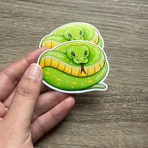 May include: Two stickers featuring a cartoon snake coiled in a circle. The snakes are bright green with yellow and white accents, and a red tongue. The stickers are held by a hand.