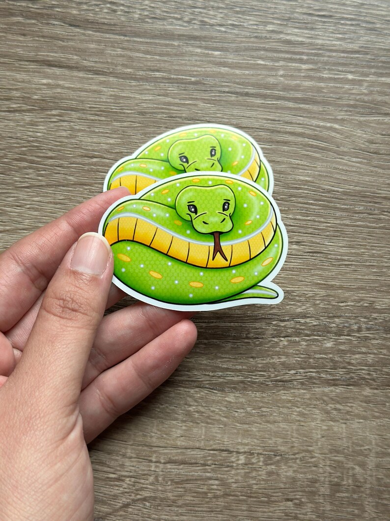 Pode incluir: Dois adesivos com uma cobra de desenho animado enrolada em um c&iacute;rculo. As cobras s&atilde;o verde brilhante com detalhes amarelos e brancos e uma l&iacute;ngua vermelha. Os adesivos s&atilde;o segurados por uma m&atilde;o.