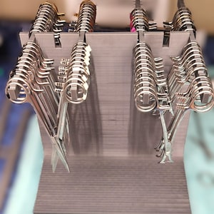 May include: A gray 3D-printed display stand featuring four vertical slots designed to hold multiple silver-colored metal surgical hemostats or scissors. The stand has a flat base and a tall back panel with notched edges to organize the instruments.