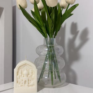 以下が含まれることがあります： 白いチューリップと緑の茎のブーケが入った透明なガラスの花瓶。花瓶の隣には、モスクまたは宮殿のような建築デザインが彫られたクリーム色のキャンドルがあります。白い表面に置かれたアイテム。