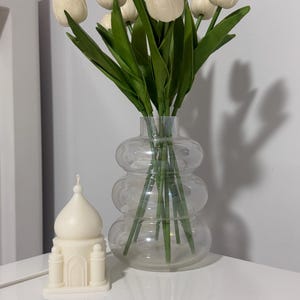 以下が含まれることがあります： ユニークなバブルデザインの透明なガラスの花瓶には、緑の茎と葉を持つクリーム色のチューリップのブーケが入っています。白いモスク型のキャンドルが花瓶の隣に置かれています。