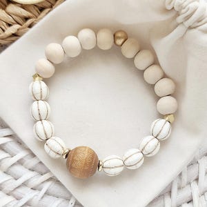 May include: A white and gold beaded bracelet with a large, brown wooden bead in the centre. The bracelet is made of round beads with a white and gold striped pattern.