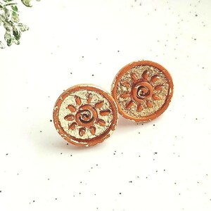 May include: A pair of round stud earrings featuring a textured copper-coloured rim and a central gold-toned inlay. Each earring displays a raised sun motif with a spiral centre and radiating rays, set against a metallic, shimmering background.