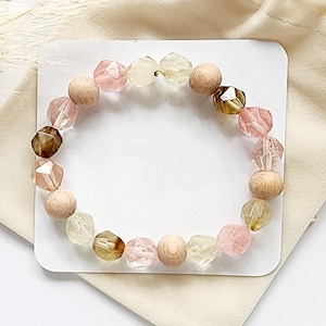 May include: A pink, brown and clear beaded bracelet with wooden beads. The bracelet is on a white card and a cream-coloured fabric background.