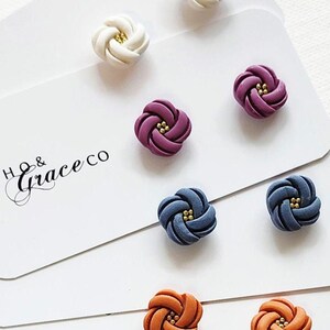 May include: Stud earrings from Boho & Grace Co. in white, purple, blue, and orange. Each earring has a knotted design with a gold centre. The earrings are presented on white cards. The brand name is visible.