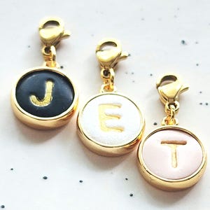 May include: Three gold-coloured initial charms with lobster clasps. Each charm features a circular design with a letter: J in gold on a black background, E in gold on white, and T in gold on pink.