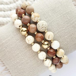 May include: A set of three bracelets featuring brown wood, white and beige stone beads, and gold accents.