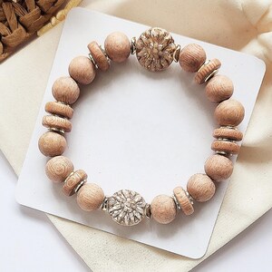 May include: A wooden beaded bracelet with two silver flower-shaped charms. The bracelet is light brown and has a natural wood finish.