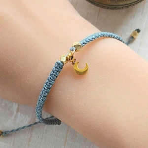 May include: A light blue macramé bracelet with gold-coloured accents. The bracelet features a small gold crescent moon charm and geometric beads. The bracelet is adjustable.