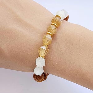 May include: A bracelet featuring gold beads and white and brown beads. The bracelet is crafted from natural materials and has a simple, elegant design.
