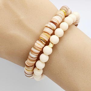 May include: Three bracelets stacked on a wrist. The top bracelet is made of small, flat, round beads in shades of cream, beige, and brown with gold accents. The middle bracelet is made of larger, round, cream-colored beads. The bottom bracelet is made of smaller, round, cream-colored beads.