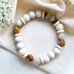May include: A bracelet with a mix of white, beige, and earth-toned beads. It features round white beads, small beige spacers, and larger, irregularly shaped beads in shades of brown, tan, and grey. The bracelet is displayed on a white cloth.