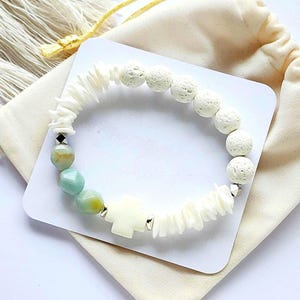 May include: A white and light blue beaded bracelet with a cross charm. The bracelet is made with lava rock, shell, and gemstone beads.