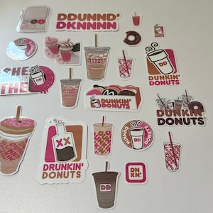 Könnte beinhalten: Eine Sammlung von Aufklebern mit Dunkin' Donuts Branding. Die Aufkleber sind rosa, weiß und schwarz mit verschiedenen Designs, darunter Donuts, Kaffeetassen und das Dunkin' Donuts Logo. Der Text auf den Aufklebern lautet "Dunkin' Donuts", "Drunkin' Donuts", "DD" und "Dunkin".