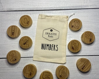 May include: A set of ten wooden number blocks with a white drawstring bag. The bag has the text "EM & KTNS Krafts NUMBERS" printed on it. Each block has a number from 0 to 9 with a circle around it.