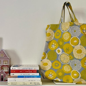 May include: A large tote bag with a repeating pattern of yellow and green floral shapes on a light blue background. The bag has matching yellow handles and is standing upright. Books and a decorative house are visible.