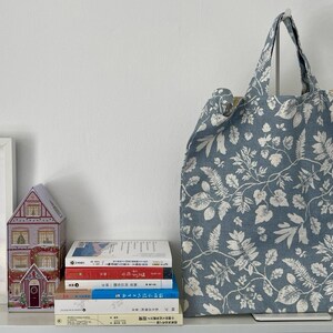 May include: A blue tote bag with a white floral pattern stands next to a stack of books and a decorative house-shaped box. The bag has two handles and a light-colored interior. The books have colorful spines.