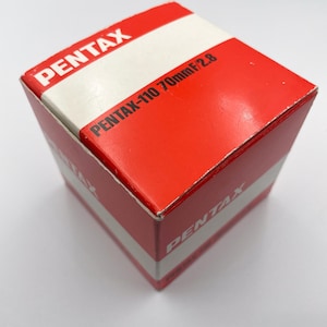 Puede incluir: Una caja de lente de cámara Pentax roja y blanca. La caja tiene la palabra "PENTAX" en letras blancas en la parte superior y lateral. El texto "PENTAX-110 70mm f/2.8" está impreso en la parte frontal de la caja.