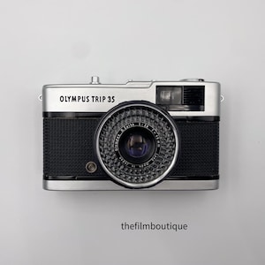 Op de afbeelding: Olympus Trip 35 camera met een zilveren bovenkant en zwarte behuizing. De camera heeft een grote lens en de tekst "OLYMPUS TRIP 35" staat op de bovenkant. Het logo van de filmboetiek staat onderaan.