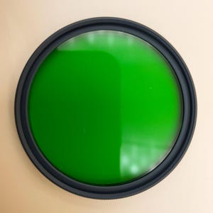 May include: A green filter for a camera lens. The filter is round and has a black plastic frame.