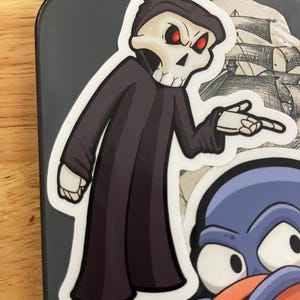 May include: A sticker featuring a cartoon Grim Reaper character with a skull face, red eyes, and a black hooded robe. The Reaper is pointing with one hand. Other stickers are visible in the background.