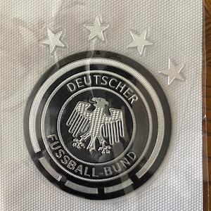 May include: A black and silver emblem with the words "DEUTSCHER FUSSBALL-BUND" and a silver eagle. The emblem is surrounded by a black border and four silver stars. The emblem is on a white textured background.