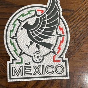 May include: A white, black, green, and red emblem featuring the word "MÉXICO" at the bottom. The emblem has a circular design with a stylized eagle and a soccer ball. The background has a textured pattern.