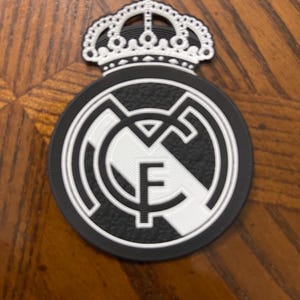 May include: A black and white emblem featuring a crown and a circular design with the letters "RMCF". The emblem is likely a team logo, possibly for a sports team. The emblem is on a wooden surface.
