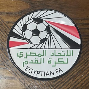 May include: A round emblem featuring a soccer ball design with black and white panels. The emblem has a red, black, and white striped section. The text "EGYPTIAN FA" is at the bottom, with Arabic text above.