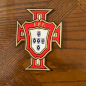 May include: A red, white, and gold emblem featuring a cross design with the letters "E.P.F." at the top. The emblem has a shield in the center with five blue and white symbols. The emblem is likely a team logo or a national symbol.