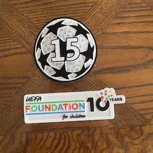 May include: Two patches on a wooden surface. The top patch is a soccer ball design with the number 15 in the center. The bottom patch reads "UEFA FOUNDATION for children" with the number 10 and the word "YEARS".