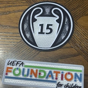 May include: Two patches on a wooden surface. One is a circular patch with a black and white design and the number 15. The other is a rectangular patch with the text "UEFA FOUNDATION for children" in various colors.