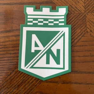 May include: A green and white patch featuring the letters "AN" inside a shield shape, with a castle-like top. The patch has a green background with white accents and a diagonal line through the letters. The patch is likely a team logo or emblem.