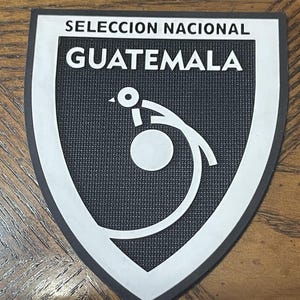 May include: A black and white shield-shaped patch with the text "SELECCION NACIONAL GUATEMALA". The patch features a white graphic of a bird and a ball. The patch has a black textured background and a white border.