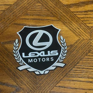 May include: A black and white Lexus Motors emblem with a shield shape, a silver laurel wreath, and the Lexus logo. The emblem is on a wooden surface with a diamond pattern.