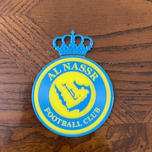 May include: A blue and yellow Al Nassr Football Club emblem. The emblem is circular with the words "AL NASSR FOOTBALL CLUB" in blue. A blue crown sits above the circle. The center of the circle is yellow with a blue design.