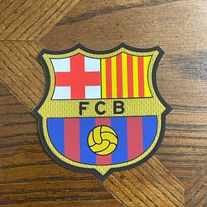 May include: A shield-shaped emblem featuring the letters "FCB" in black. The top half displays a white cross on a red background and yellow and red vertical stripes. The bottom half has blue and red vertical stripes with a yellow soccer ball.