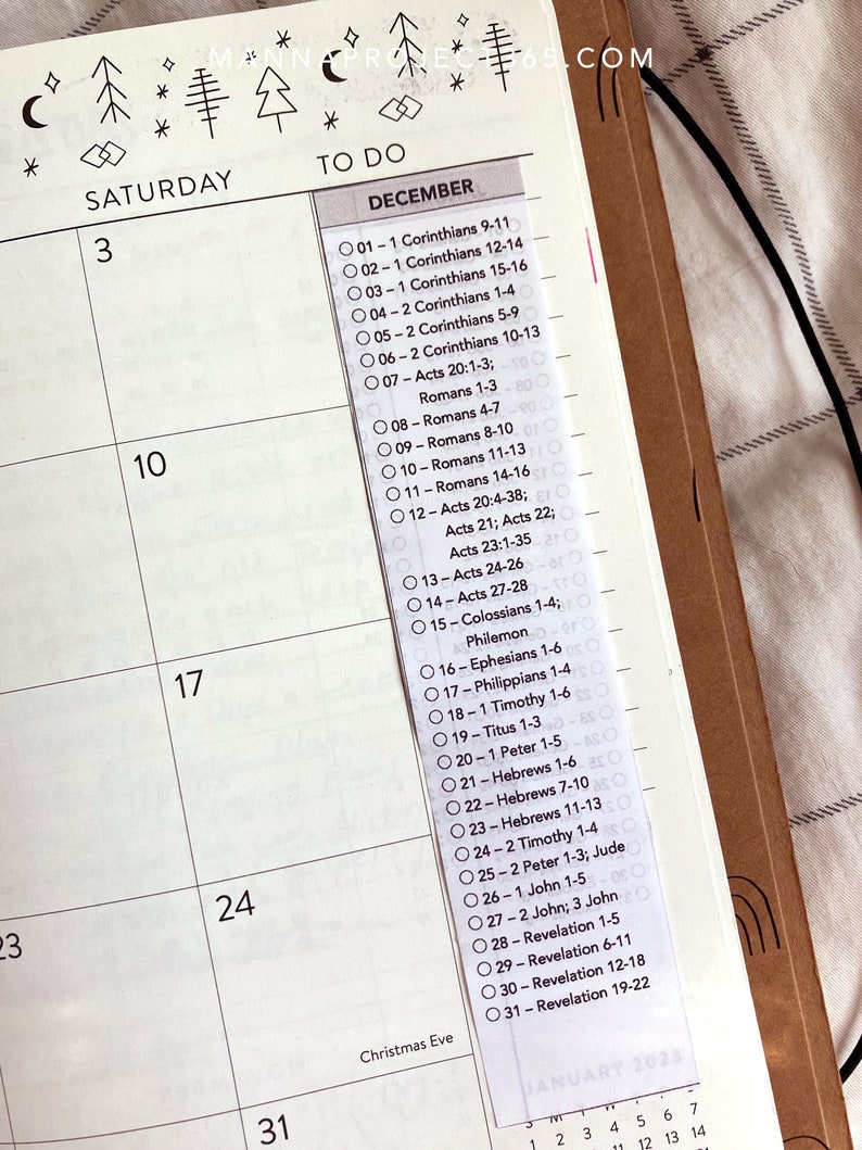 May include: A planner page with a December calendar and a list of Bible verses. The top of the page has decorative tree and star illustrations. The text "TO DO" is above the list of verses. The planner is open to the week of December 3rd.